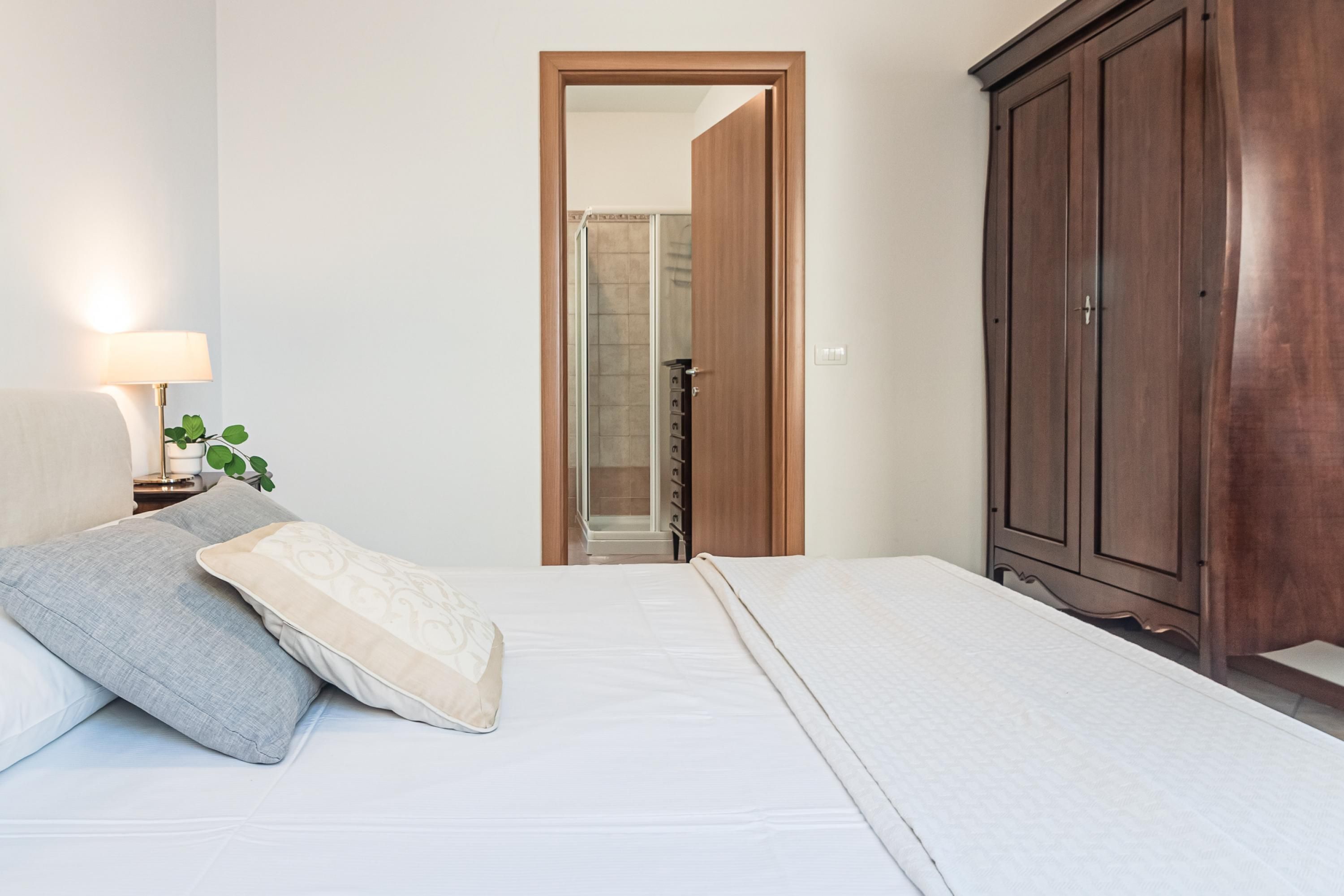 A bedroom with a bed, a nightstand with a lamp, a wardrobe, and a bathroom visible through an open door.