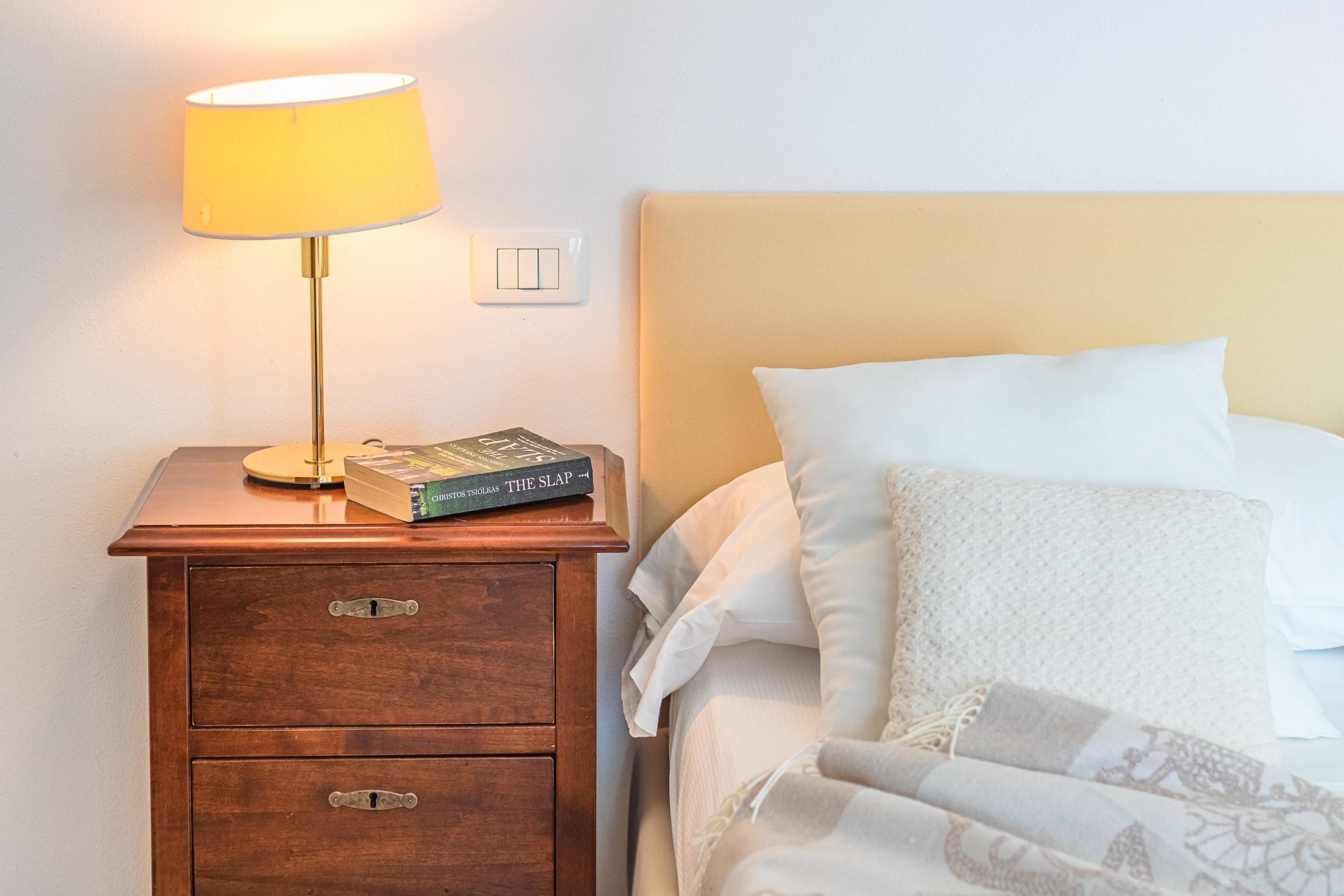A neatly made bed with white linens and a patterned throw blanket, a bedside lamp, and a nightstand with a book titled "The Slap" and a power outlet.
