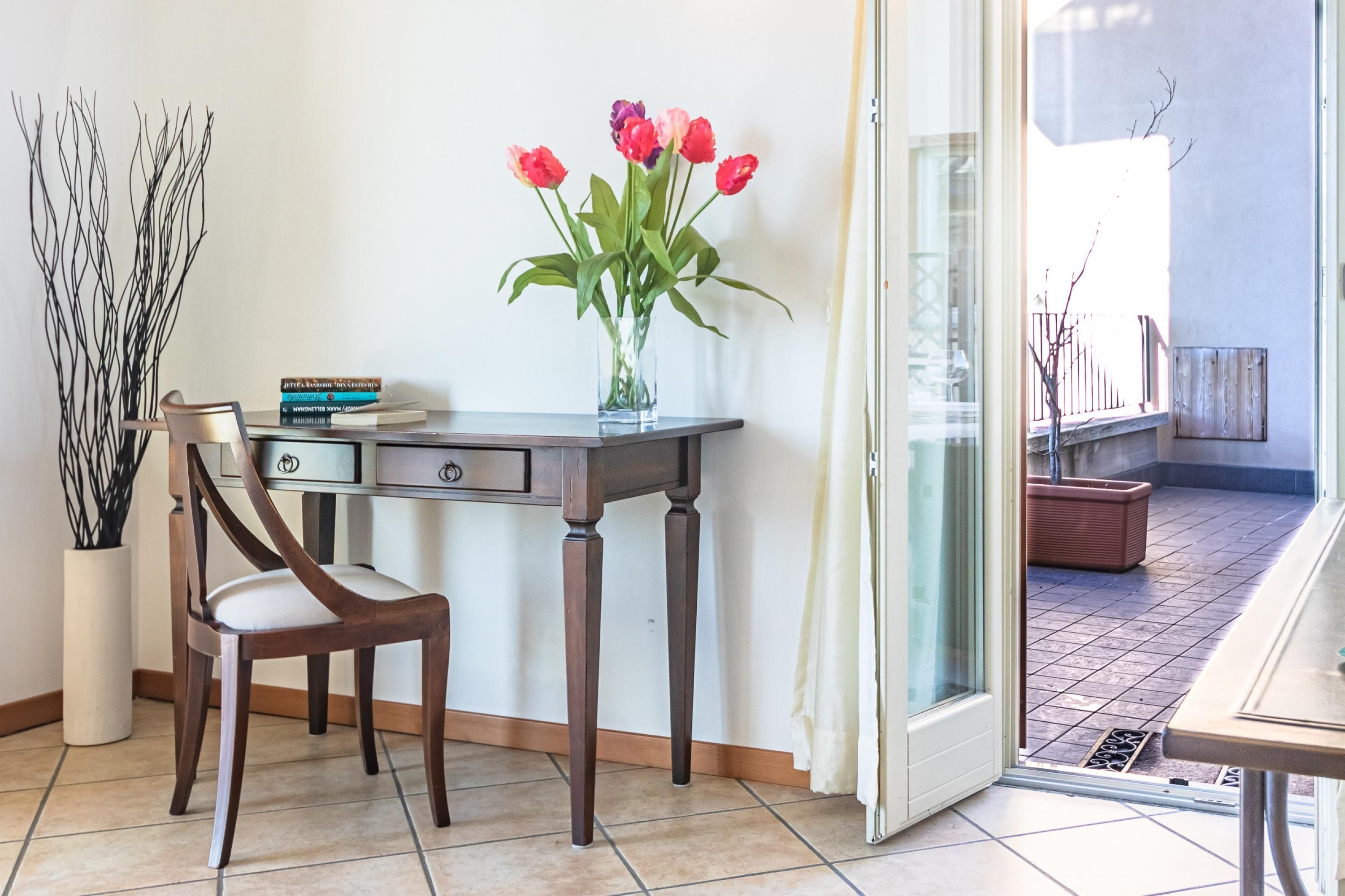 A room with a wooden desk, a chair, a vase with red flowers, and a view of the outdoor area through a glass door.
