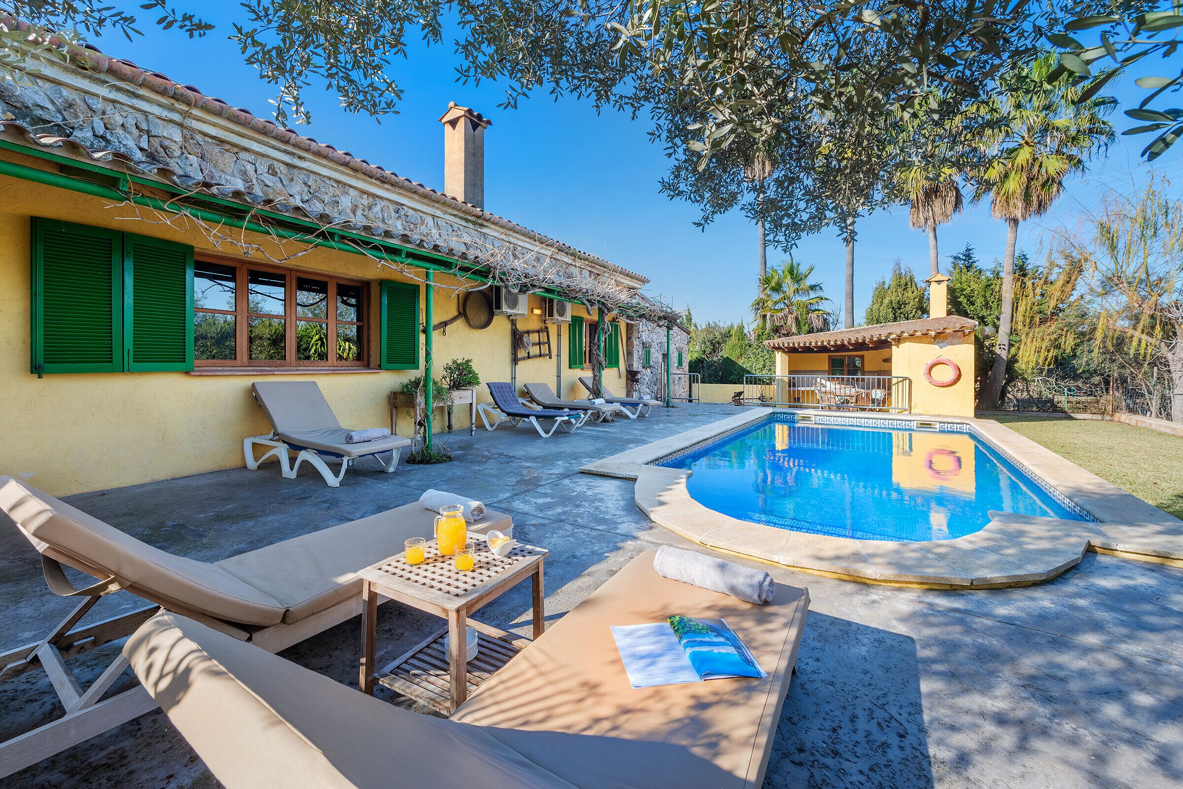 Zona de piscina con sillones reclinables, una mesa con bebidas y un edificio pequeño con chimenea.