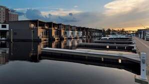 Een jachthaven met aangemeerde boten, een rij gebouwen met balkons en een rustig wateroppervlak dat de gebouwen weerspiegelt.