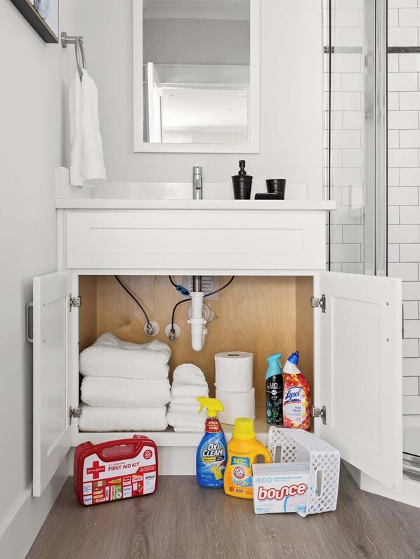 Baño con lavamanos blanco, espejo encima y un armario debajo. Se ven toallas, un botiquín, productos de limpieza y una caja de pañuelos.