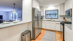 Una cocina moderna con armarios blancos, electrodomésticos de acero inoxidable, un ventanal y piso de baldosas.