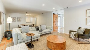 Una sala de estar moderna con sofá, una mesa de centro y una silla. Hay una puerta que conduce a otra habitación y una ventana con persianas.