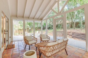 Eine überdachte Terrasse mit Korbmöbeln, Fliesenboden und großen Fenstern mit Blick auf den Garten.