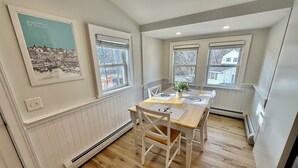 Un coin repas avec une table en bois, des chaises blanches et une plante en pot. Il y a trois fenêtres équipées de stores et un tableau encadré au mur.