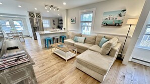 Un salon moderne avec un canapé d’angle beige, une table basse et un plancher en bois. On y trouve trois tabourets de bar bleus, un coin cuisine avec une grande fenêtre et un tableau représentant un camion sur le mur.