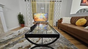 Een woonkamer met een salontafel met glazen blad, een bruine bank met gele kussens, een televisie en een kamerbreed tapijt met patroon.