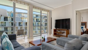 A modern living room with grey sofas, a wooden coffee table, and a flat-screen TV. Large windows offer views of a multi-story building. - Contemporary Cape Town Gem- No Loadshedding (Cape Town)