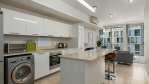 A modern kitchen with white cabinets, a built-in washing machine, and a large island with bar stools. - Contemporary Cape Town Gem- No Loadshedding (Cape Town)