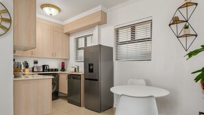 A modern kitchen with light wood cabinets, stainless steel appliances, a white dining table, and a geometric wall decoration. - Chic Beachfront Oasis - Balcony - Netflix (Cape Town)