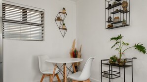 A dining area with a round table, two white chairs, a black metal shelf with decorative items, and a window with blinds. - Chic Beachfront Oasis - Balcony - Netflix (Cape Town)