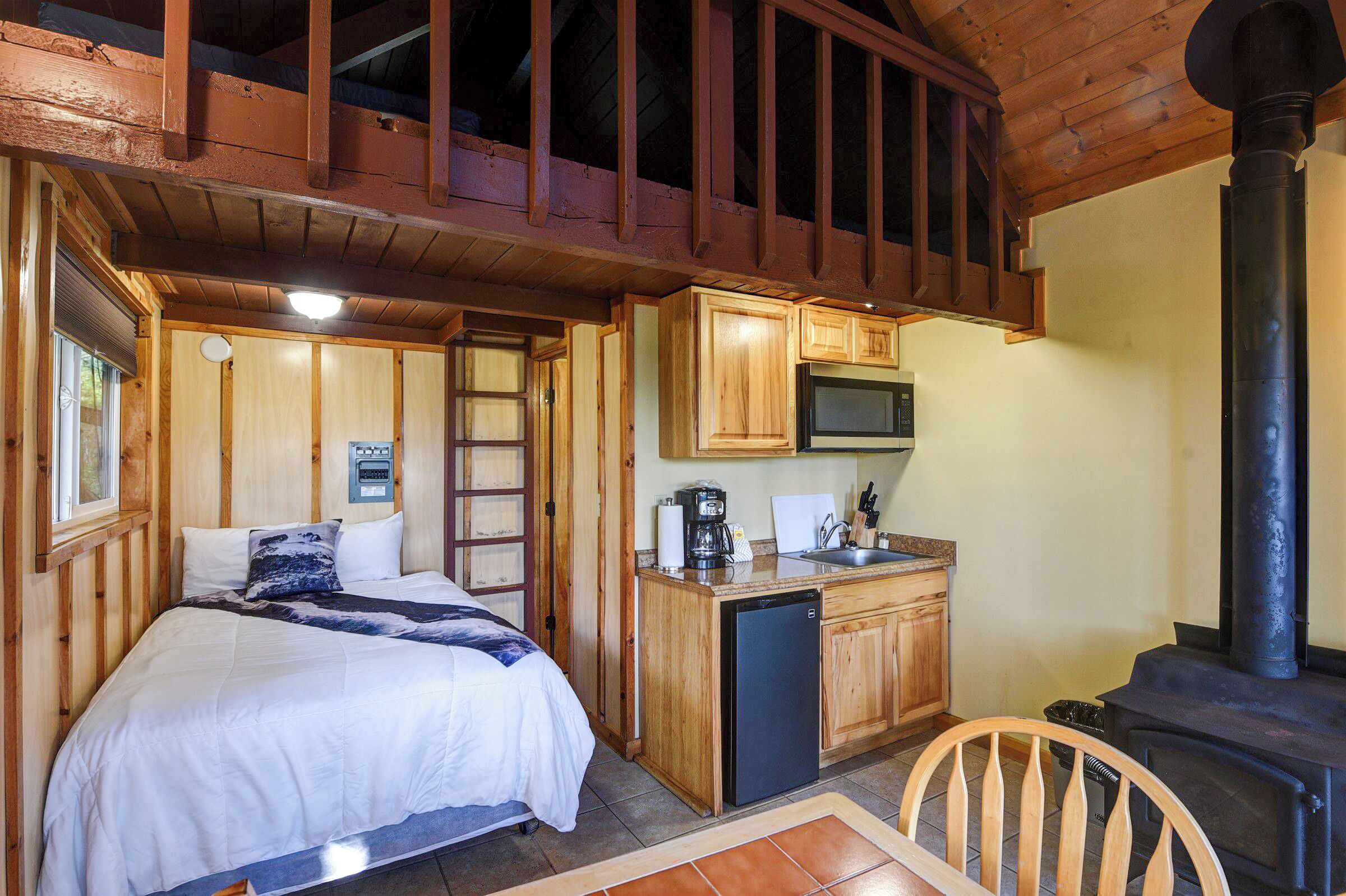 A compact room with a bed, wooden cabinets, a microwave, and a wood-burning stove.
