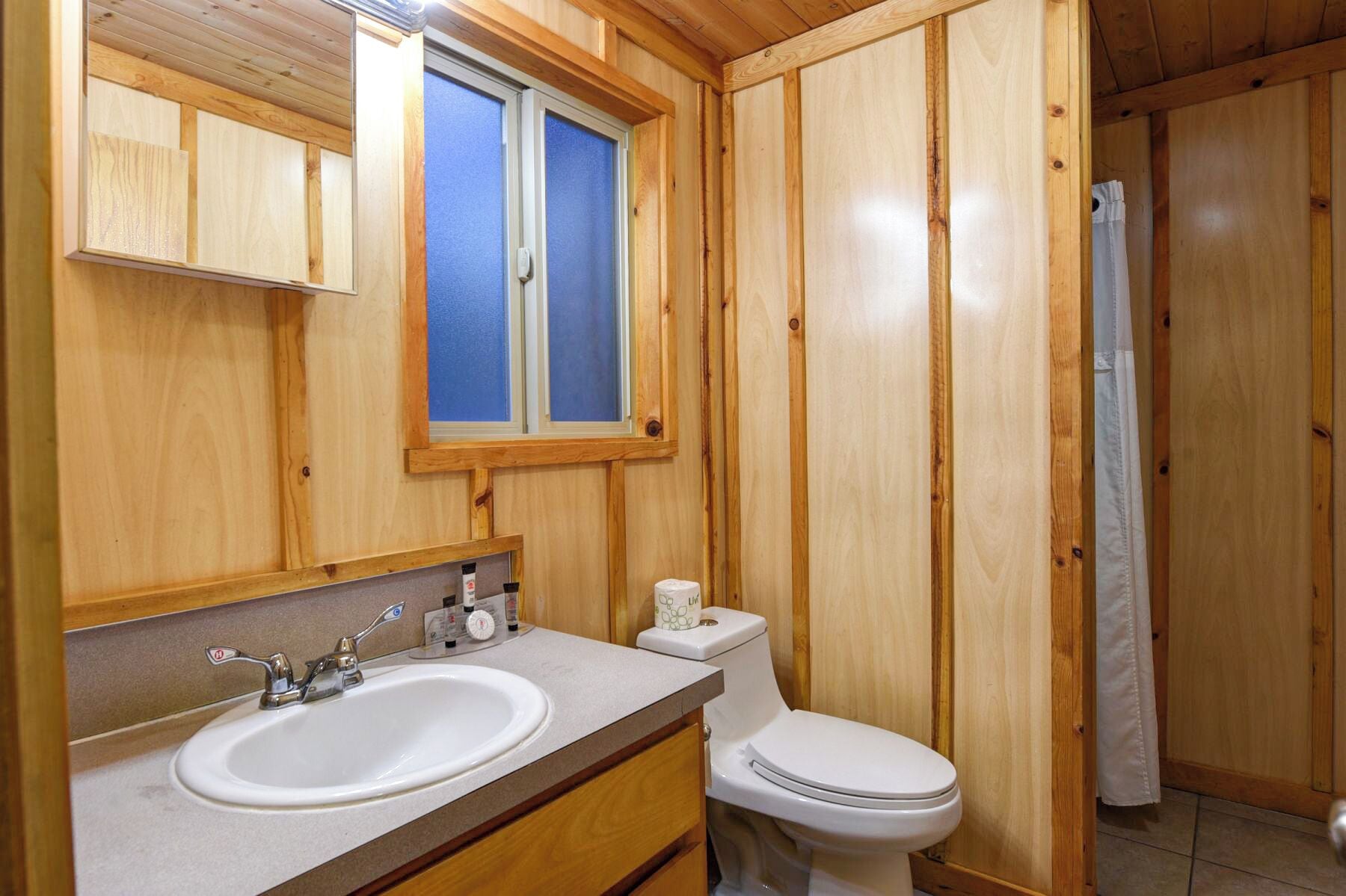 A wooden bathroom with a double sink, toilet, and a window.