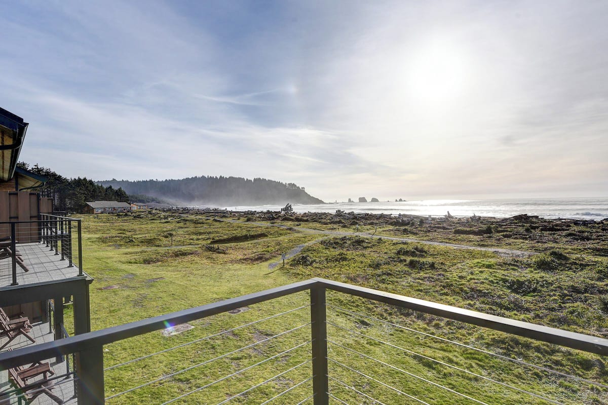 A coastal view from a balcony with a grassy area, a wooden deck, and a distant forested hill.