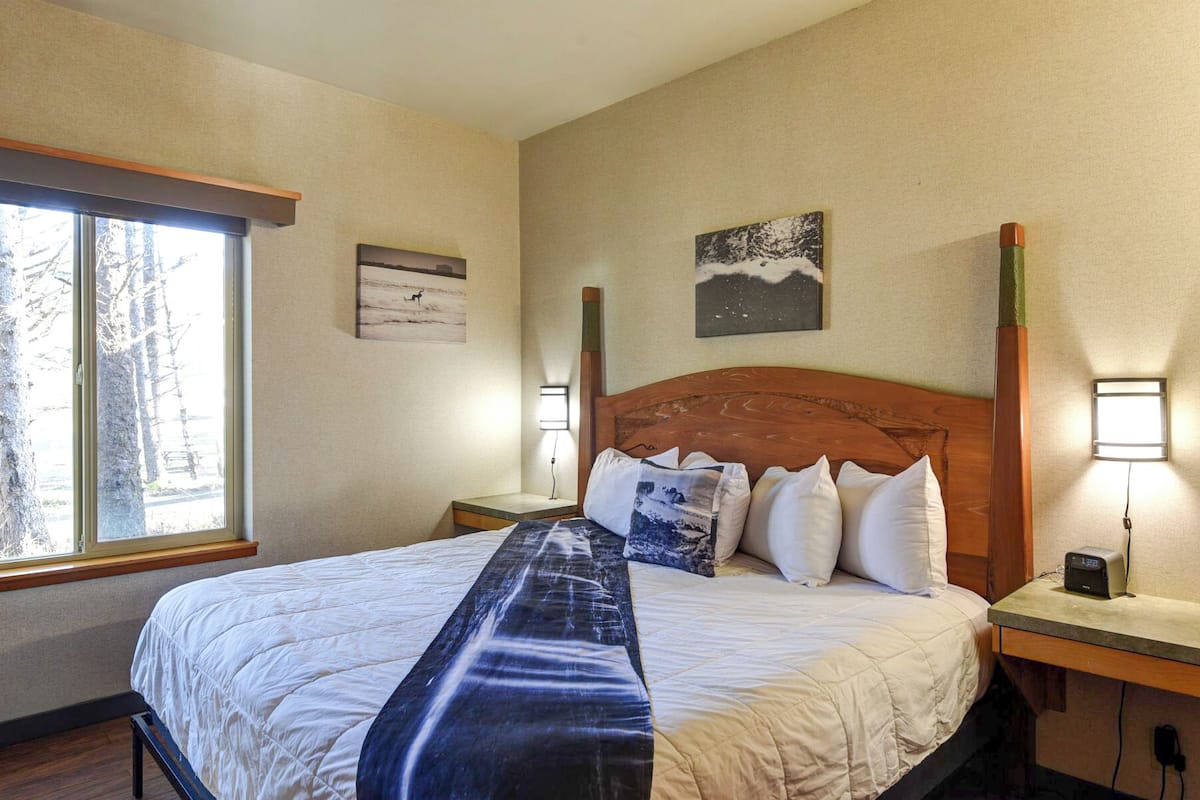 A hotel room with a large bed, two bedside tables, a window with a view of trees, and two wall-mounted lamps.