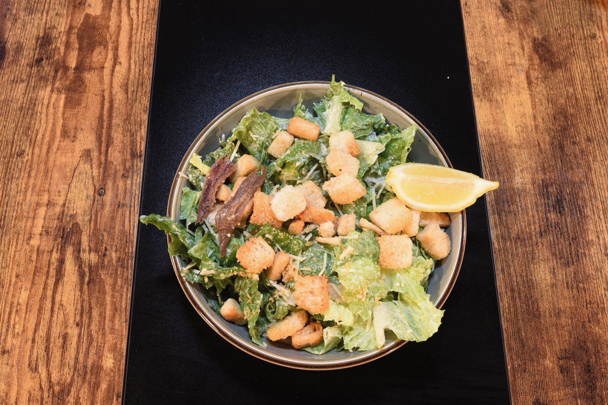 A bowl of salad with croutons and a lemon wedge on a wooden table.