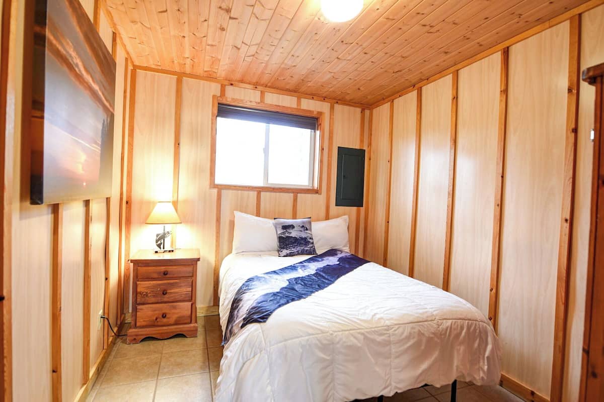 A bedroom with wooden walls, a bed with white and blue bedding, a nightstand with a lamp, and a window with blinds.