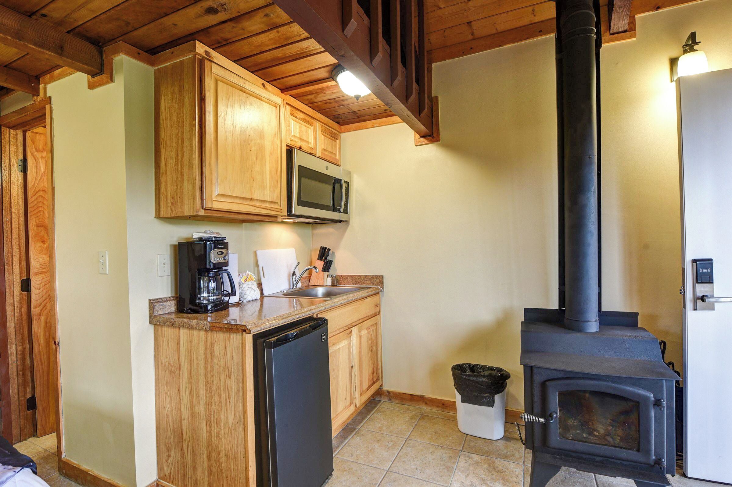 Una cocina con gabinetes de madera, microondas, cafetera, pileta, lavavajillas y estufa a leña.