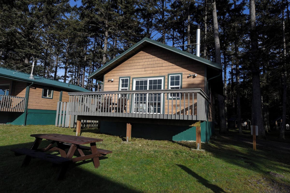 A cabin with a deck, a picnic table, and a forest backdrop.
