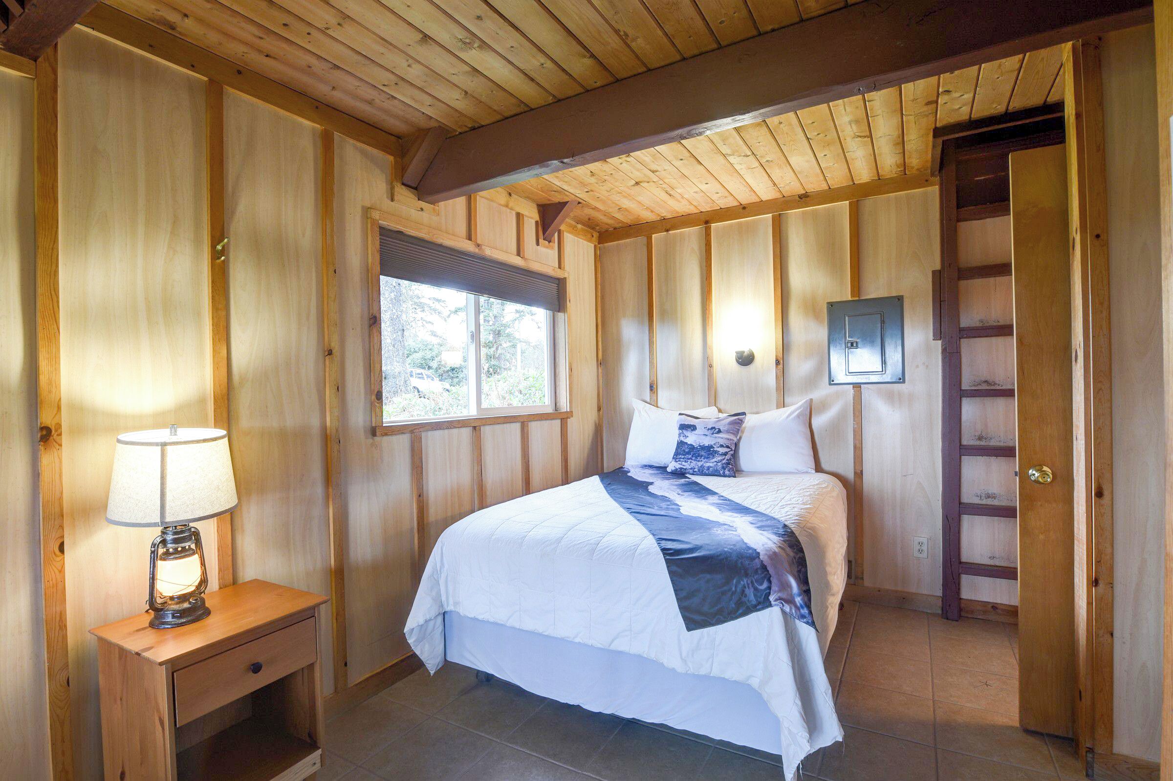 A wooden-paneled bedroom with a bed, a nightstand, a lamp, a window, and a door with a ladder.