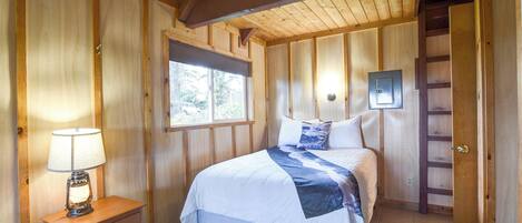 A wooden-paneled bedroom with a bed, a nightstand, a lamp, a window, and a door with a ladder.