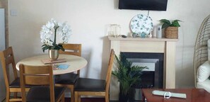 A cozy room with a round dining table, two wooden chairs, a vase with white flowers, a fireplace, and a television mounted on the wall. - 2 Bed Chalet Sleeps 5 - Pets, Parking, Near Beach (Great Yarmouth)