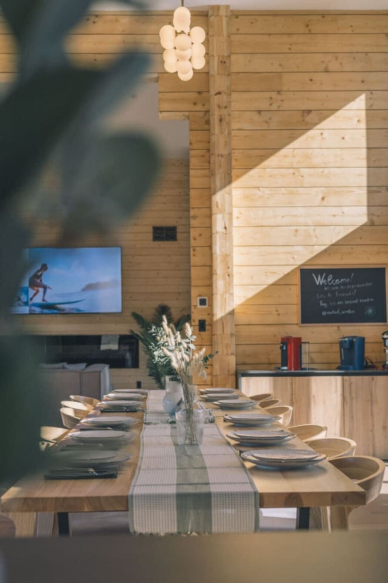 A wooden interior with a long dining table set for a meal, a television on the wall, and a chalkboard with a welcome message.