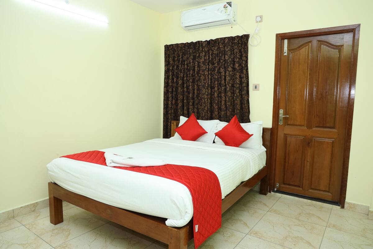 A neatly made bed with a red and white bedspread, a wooden headboard, and a red pillow. There is a wooden door to the right and an air conditioning unit on the wall above the bed.