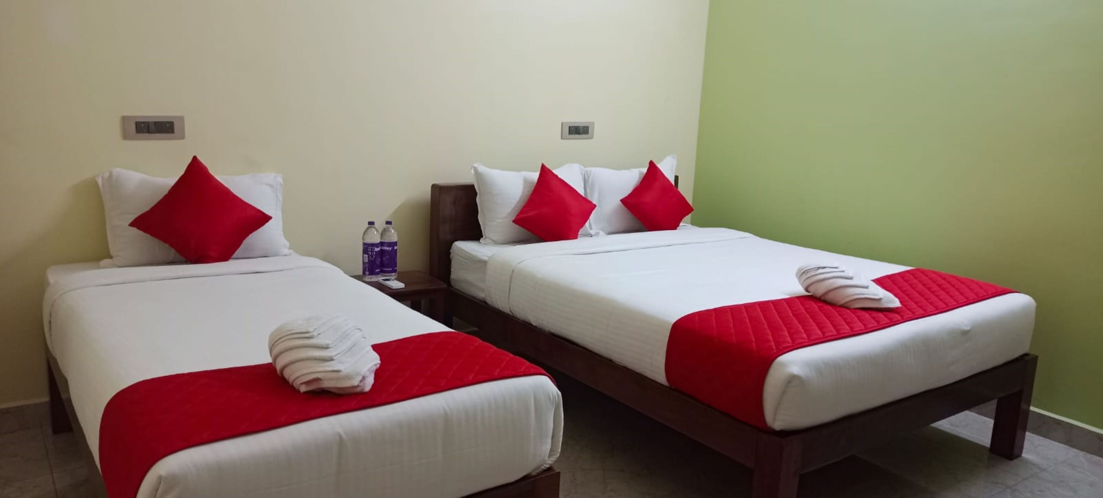Dos camas individuales con sábanas blancas y almohadas rojas, una mesita de noche con botellas de agua y una pared verde.