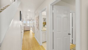 Un pasillo con piso de madera, paredes blancas y una puerta de color blanco. Se observa una escalera a la izquierda y una cama con cabecera al fondo.