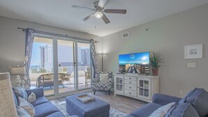 Ein Wohnzimmer mit einer Sofagarnitur, einem Fernseher und Blick auf einen Balkon mit Gartenmöbeln.