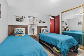 A room with two beds, each with a blue bedspread and pillows with 'TO THE BEACH' text, a wooden headboard, a nightstand with a lamp, and a red door. - Massive 3 bedroom with Pool (Palmas del Mar)
