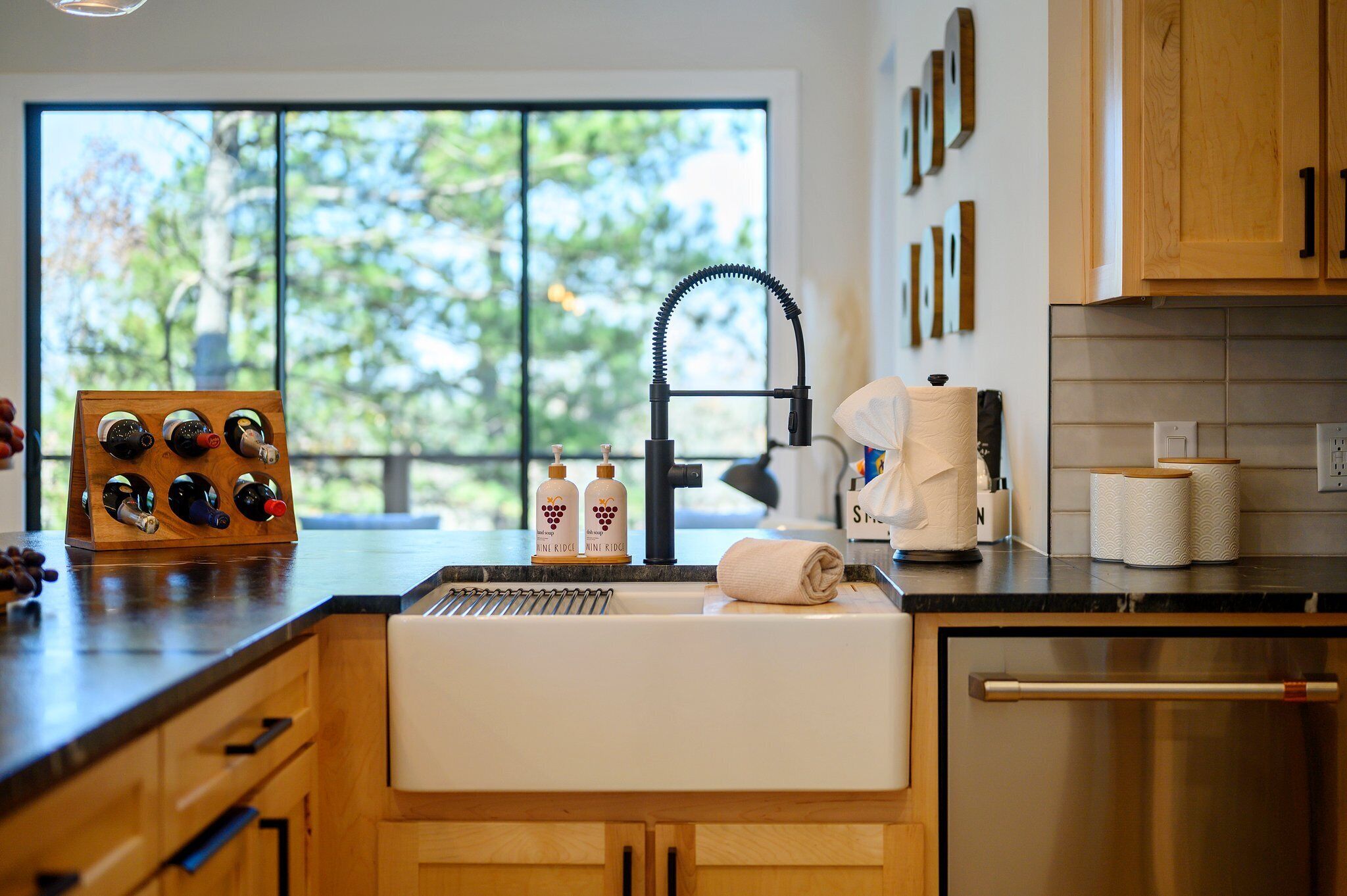 A modern kitchen with a built-in sink, wooden cabinets, a wine rack, and a view of trees through a large window.