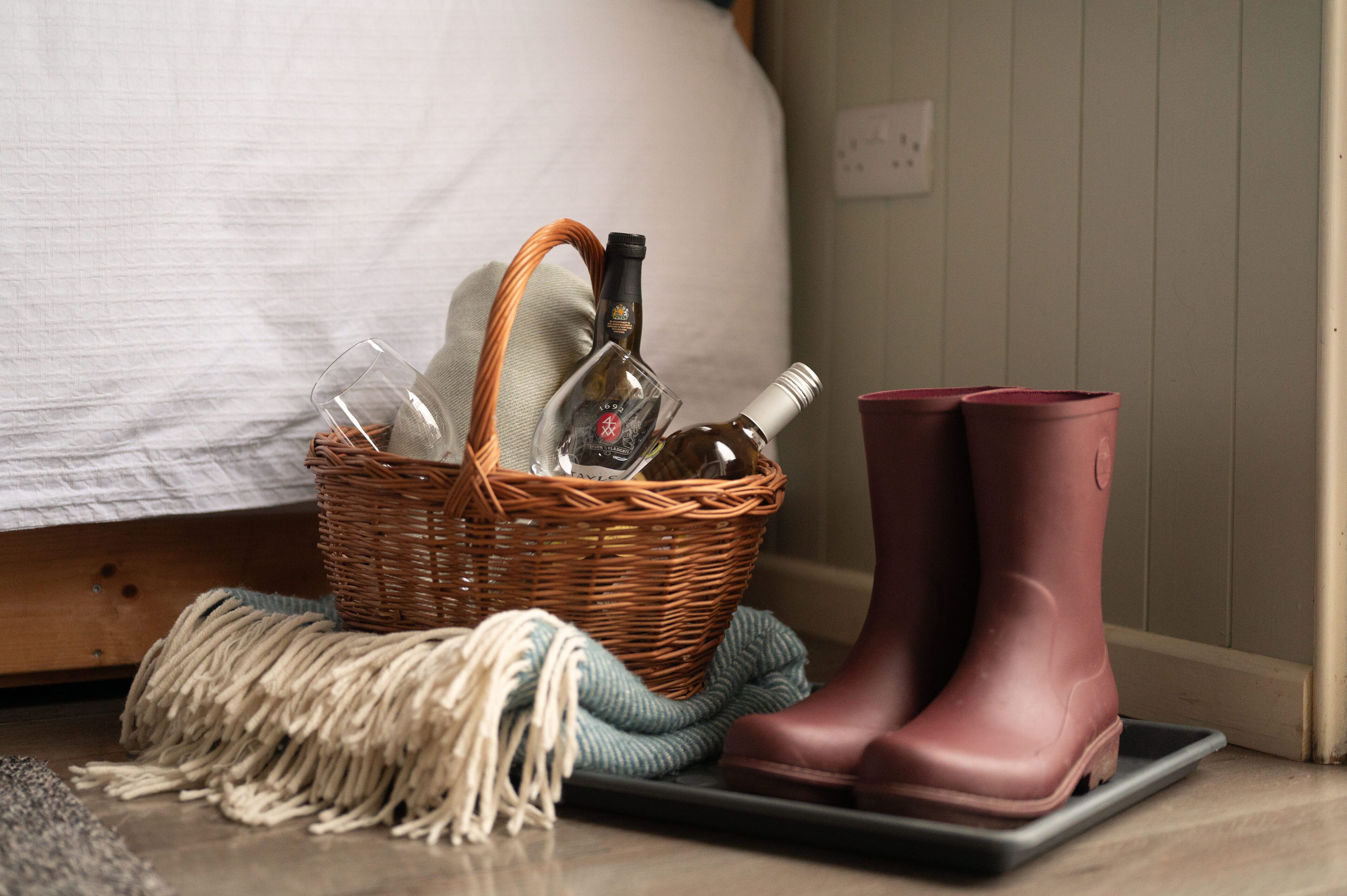 A wicker basket with bottles and a glass on a bed, next to a pair of pink rubber boots and a green rope.