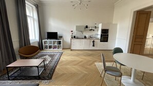 Una sala moderna con sofá, mesa de centro y televisor. Cuenta con una cocina equipada con microondas, horno y armarios. El ambiente tiene piso de madera y una lámpara de araña.