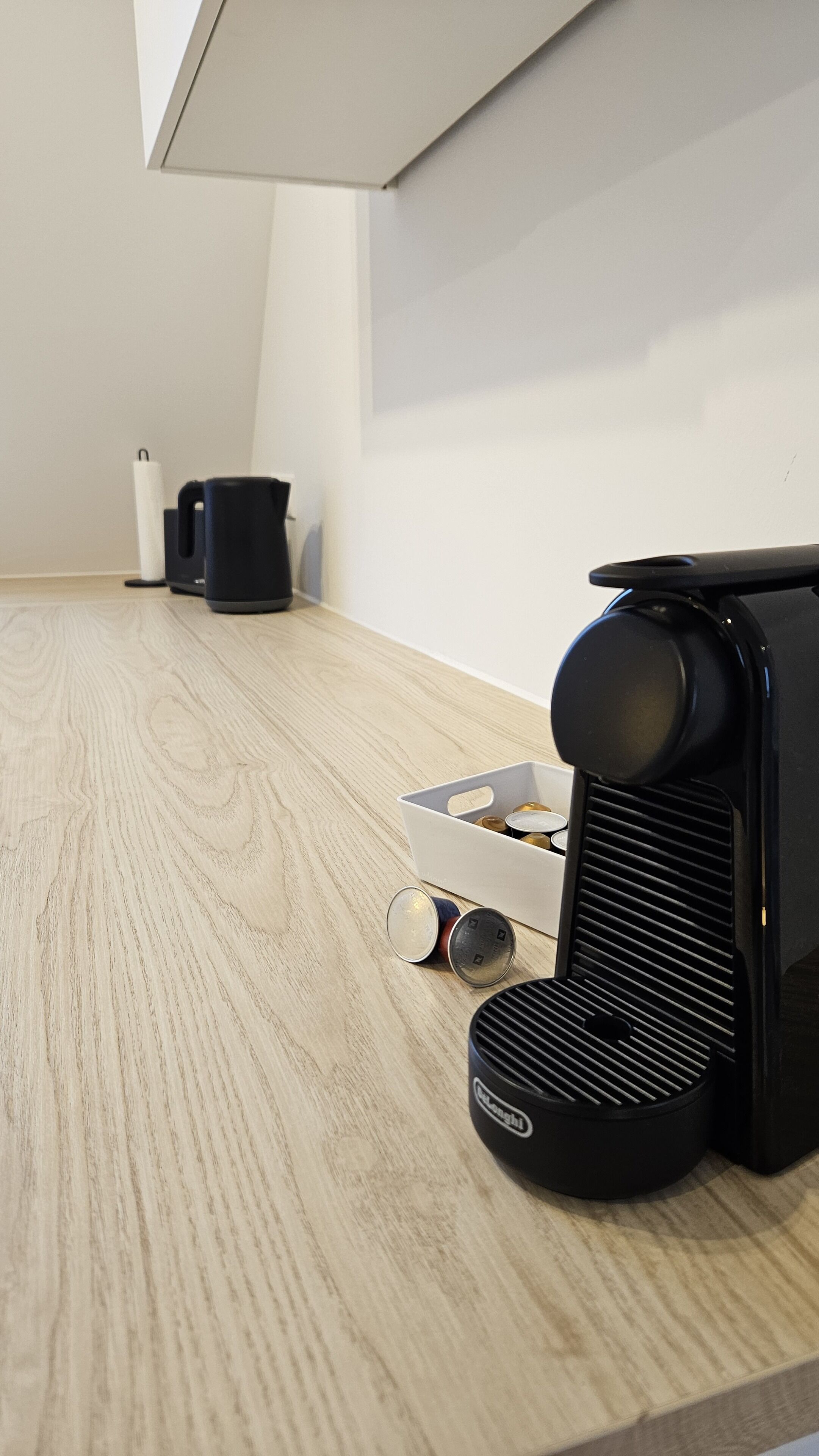 A kitchen with a coffee machine, a trash bin, and a container with cans on a wooden floor.