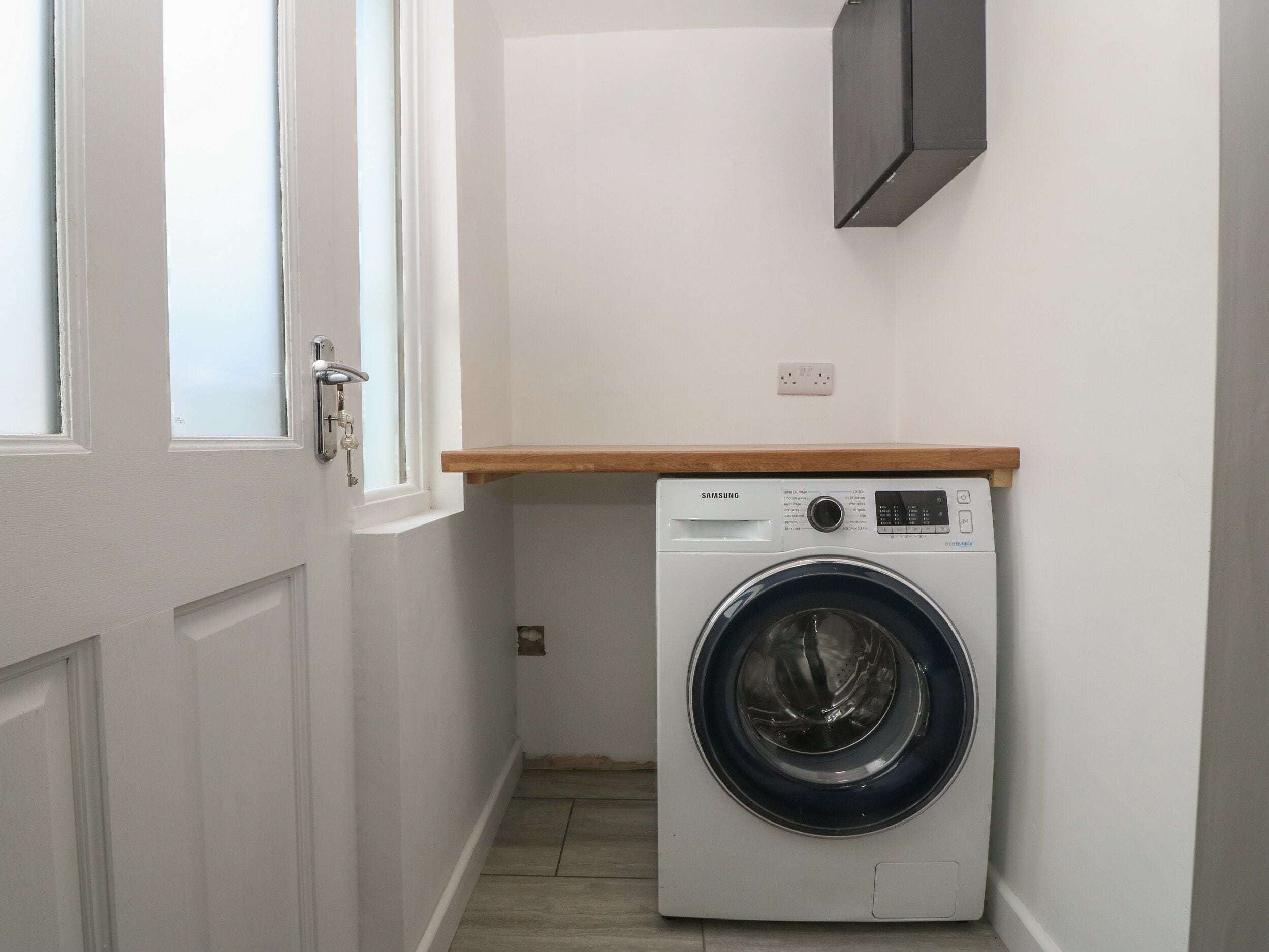 A compact laundry room with a front-loading washing machine, a wooden shelf above, and a white door.