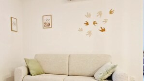Una sala de estar con un sofá color beige, dos cojines, una mesita redonda con una vela de vidrio, y detalles decorativos en las paredes, incluyendo cuadros enmarcados y adhesivos con forma de hojas.