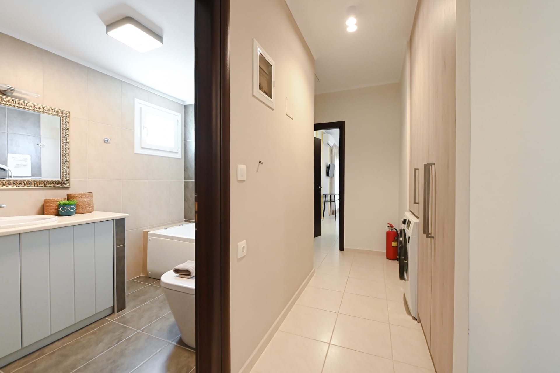 A modern bathroom with a bathtub, toilet, and vanity. A hallway leads to a living area with a fire extinguisher and a door.