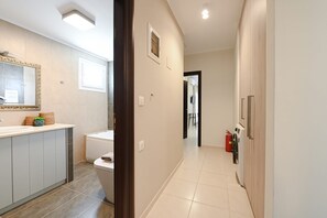 Une salle de bain moderne équipée d’une baignoire, de toilettes et d’un meuble-lavabo. Un couloir mène à un espace de vie où se trouvent un extincteur et une porte.