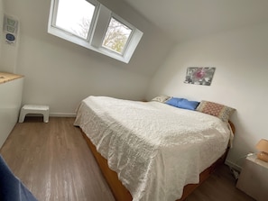 Une chambre à coucher comprenant un lit, un petit tabouret, une table de chevet, une fenêtre et un thermostat fixé au mur.