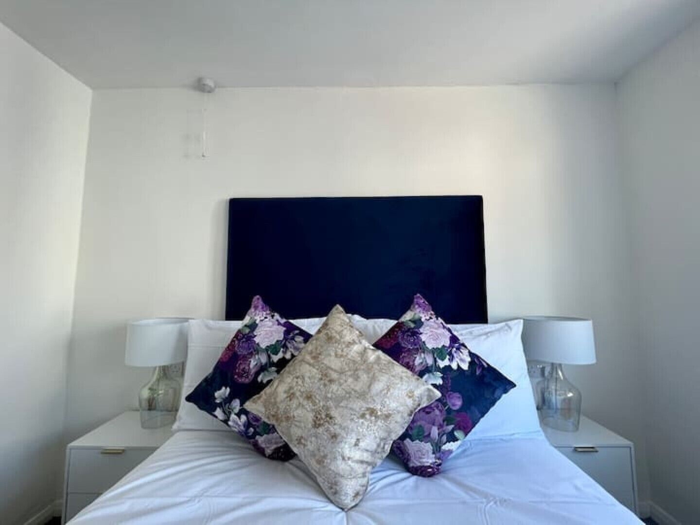 Ein ordentlich bezogenes Bett mit floralen und strukturierten Kissen, zwei Nachttischlampen und einem dunklen Kopfteil in einem schlichten Zimmer.