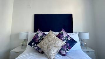 Ein ordentlich bezogenes Bett mit floralen und strukturierten Kissen, zwei Nachttischlampen und einem dunklen Kopfteil in einem schlichten Zimmer.