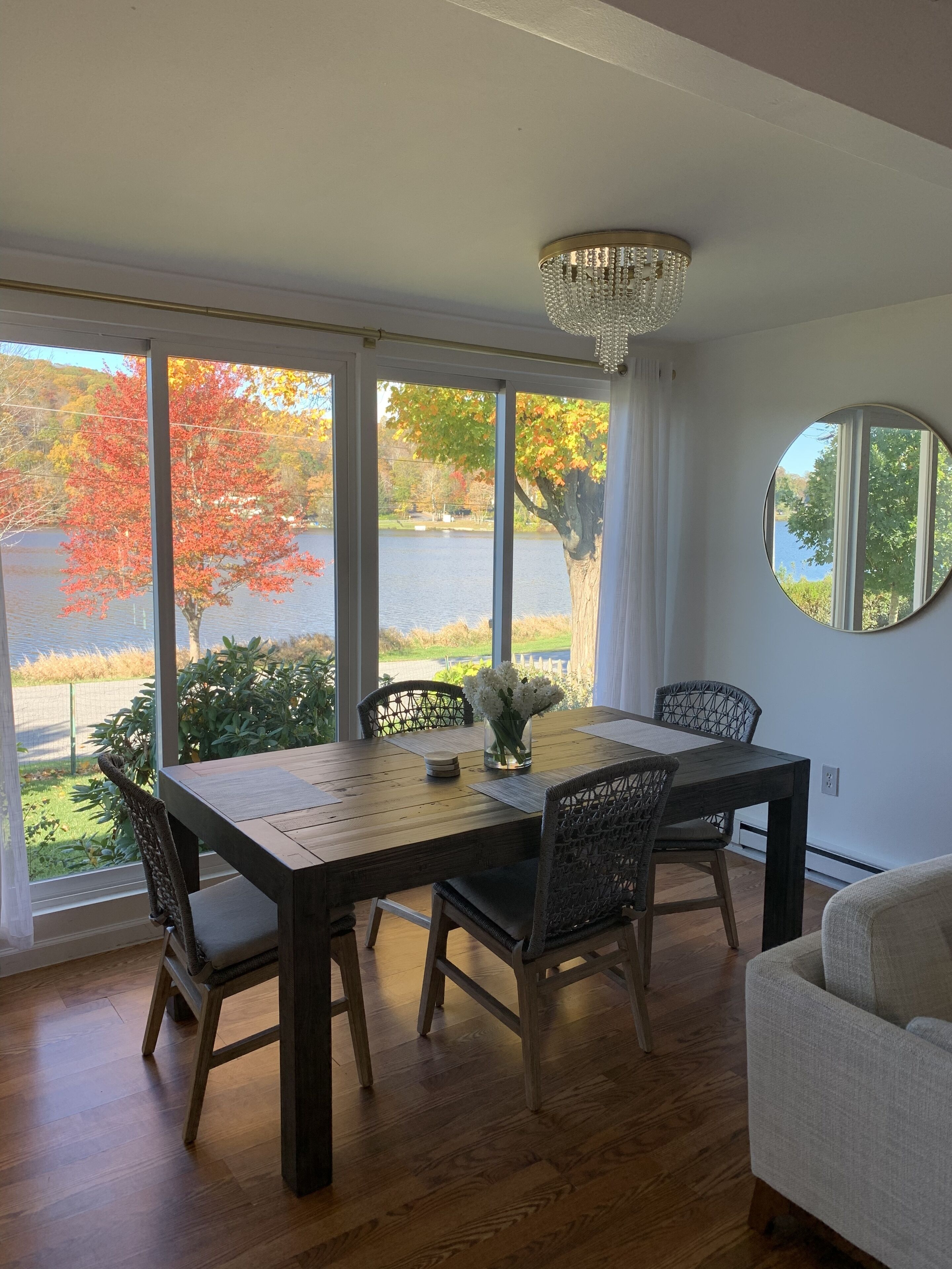 A dining room with a wooden table and chairs, large windows with a view of trees and a lake, and a chandelier.