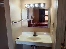 Un baño de hotel con un lavabo blanco, un espejo con iluminación y una cama visible en el reflejo.