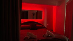 Una habitación tenuemente iluminada con un tono rojizo, que incluye una cama con toallas enrolladas, un cabecero oscuro y un perchero de pared.