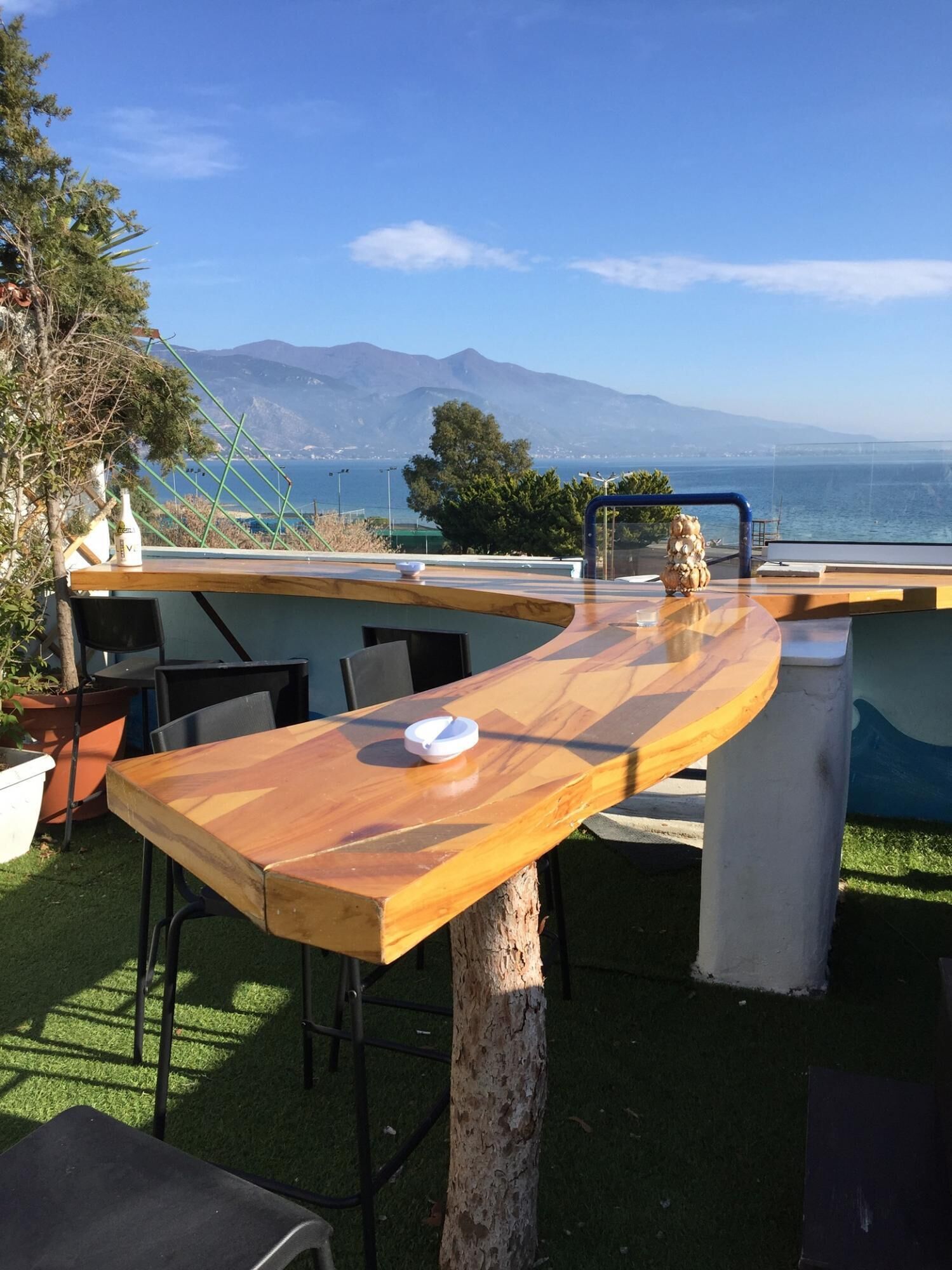 An outdoor bar area with a wooden table, chairs, and a view of the sea and mountains.