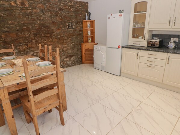A kitchen with a stone wall, wooden chairs, a dining table set, a washing machine, and cabinets. - The Granary (Kilgetty)