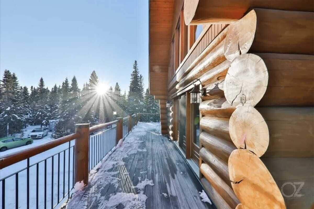 A wooden deck with snow-covered ground, a wooden railing, and a view of snow-covered trees and a car in the background.
